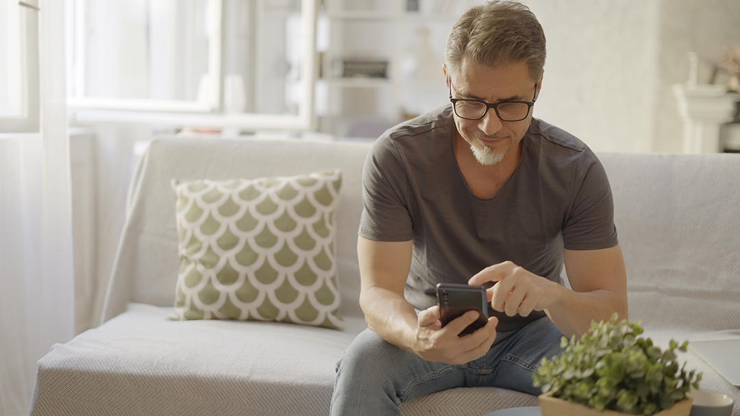 Mann von vorne sitzt auf dem Sofa und bedient sein Handy 